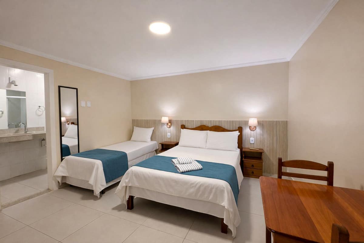 A hotel room with two single beds, a wooden table, a chair, a mirror, and a bathroom with a sink and mirror.