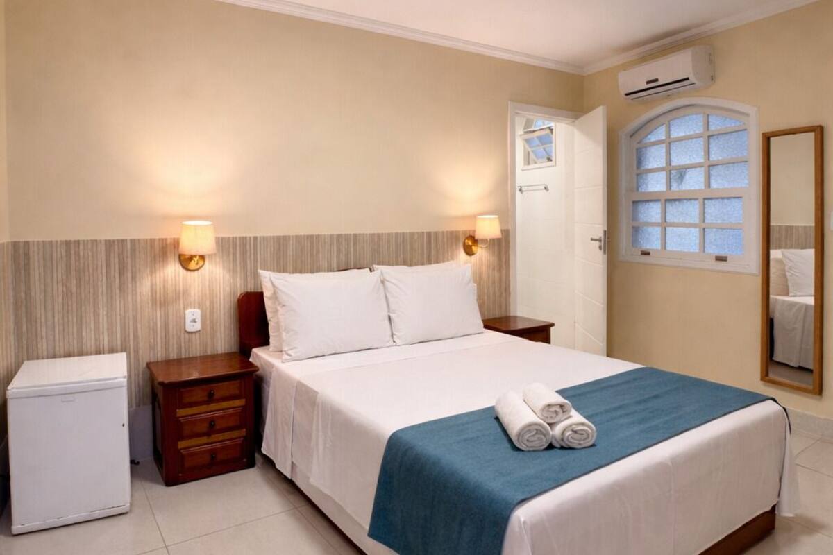 A hotel room with a bed, bedside tables, a wardrobe, an air conditioning unit, a mirror, and a window.