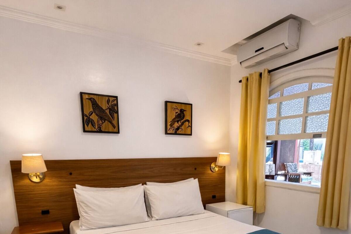 A neatly made bed with a teal bedspread and white pillows, two wall-mounted lamps, and two framed artworks on the wall.