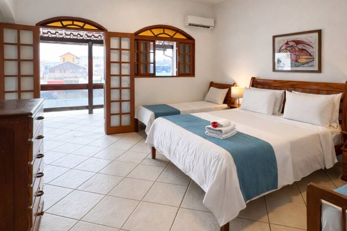 A hotel room with two beds, a wooden dresser, a desk, and a large window with a view of buildings.