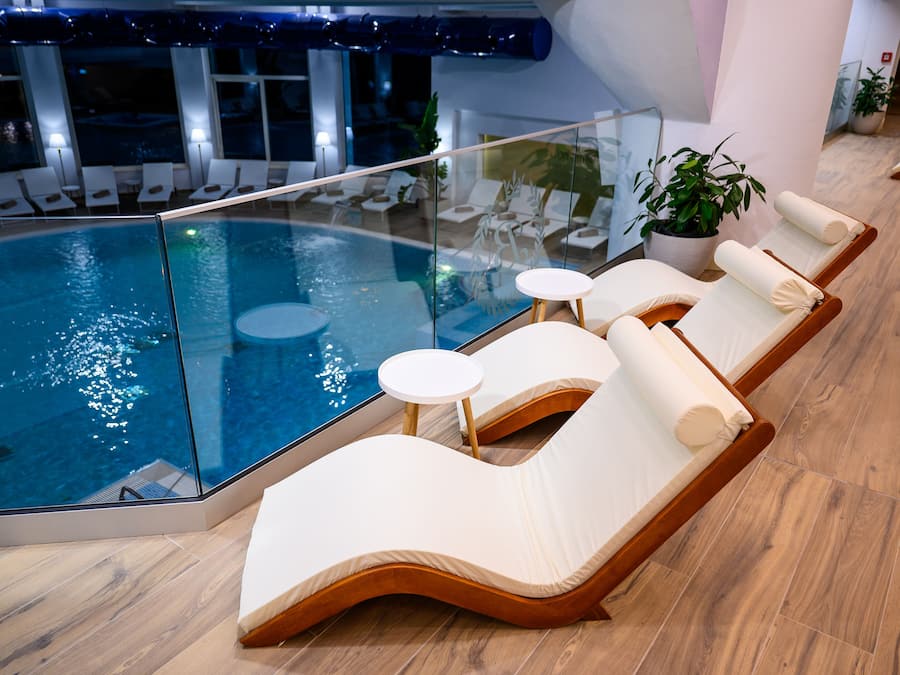 Un espace piscine intérieur moderne, doté de murs en verre, de chaises longues en bois et de petites tables rondes.