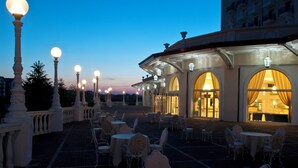 An outdoor patio with tables and chairs, a building with arched windows, and decorative street lamps. - Grand Hotel Italia (Cluj-Napoca)