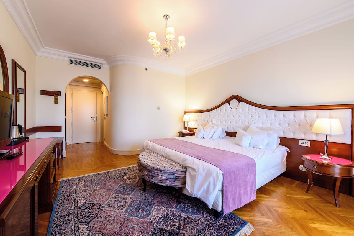 A hotel room with a large bed, a wooden nightstand, a desk with a chair, a patterned rug, and a chandelier.