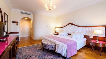 A hotel room with a large bed, a wooden nightstand, a desk with a chair, a patterned rug, and a chandelier.