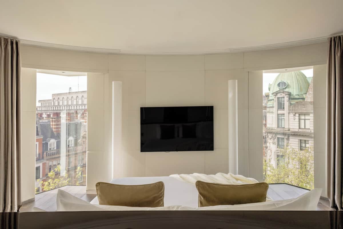 A hotel room with a bed, a flat-screen TV mounted on the wall, and large windows offering a city view.
