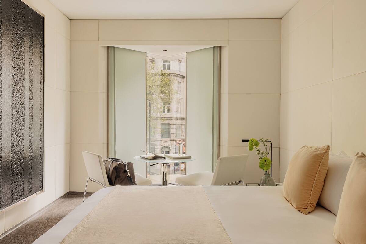 A modern hotel room with a large bed, a small table, two chairs, a view of a cityscape through a window, and a decorative wall piece.