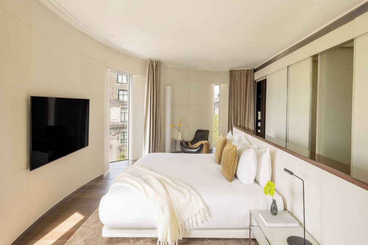 A modern hotel room with a large bed, a flat-screen TV mounted on the wall, a bedside table with a lamp, and a view of the city through large windows.
