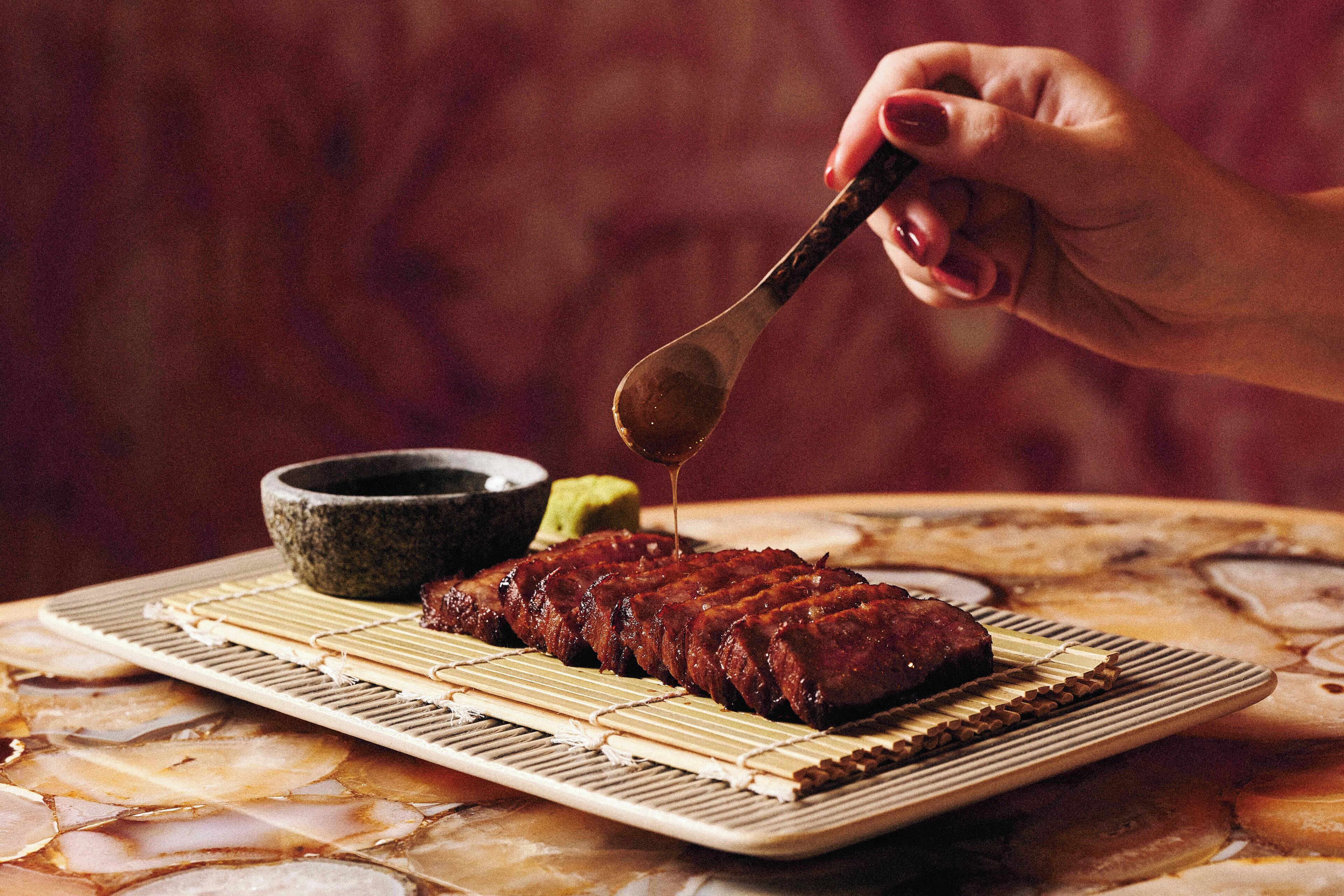 A hand pouring sauce over a dish of barbecued meat on a plate.