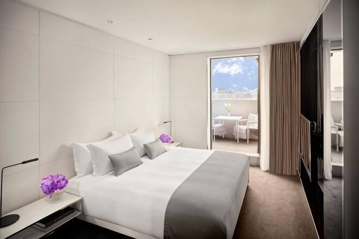 A modern hotel room with a large bed, a flat-screen TV, a bedside table, and a view of the outdoor terrace.