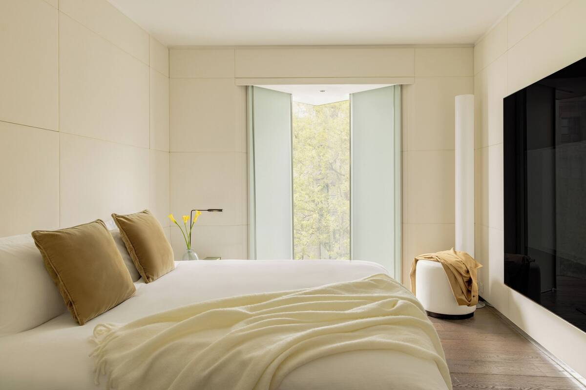 A modern bedroom with a large bed, a window with a view of trees, and a black screen on the wall.