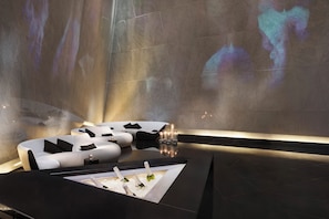 Un intérieur moderne avec un canapé aux lignes courbes, une petite table sur laquelle sont posées des bouteilles, et une projection sur le mur.