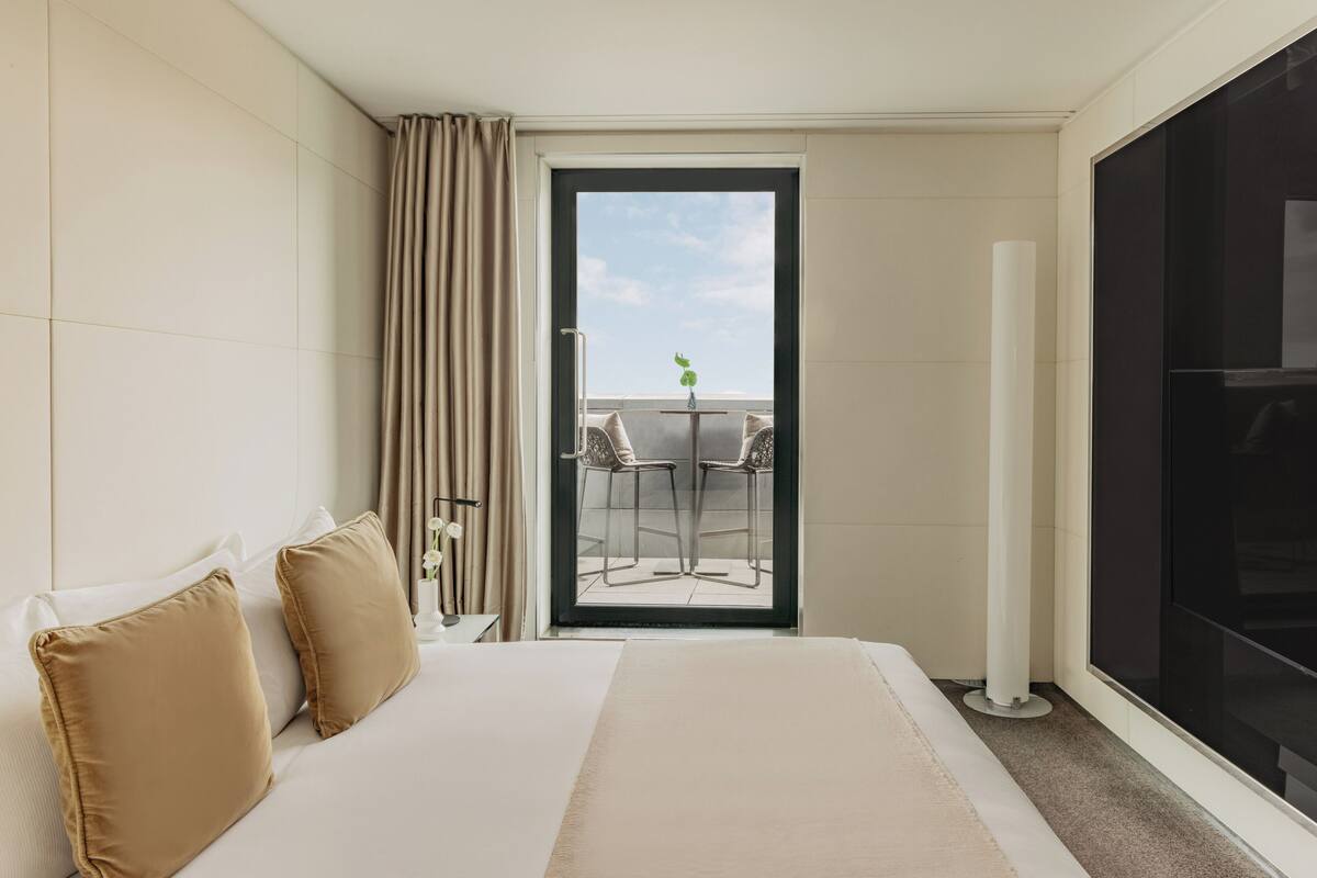 A hotel room with a bed, two pillows, a nightstand, a window with curtains, and a view of a balcony with chairs and a table.