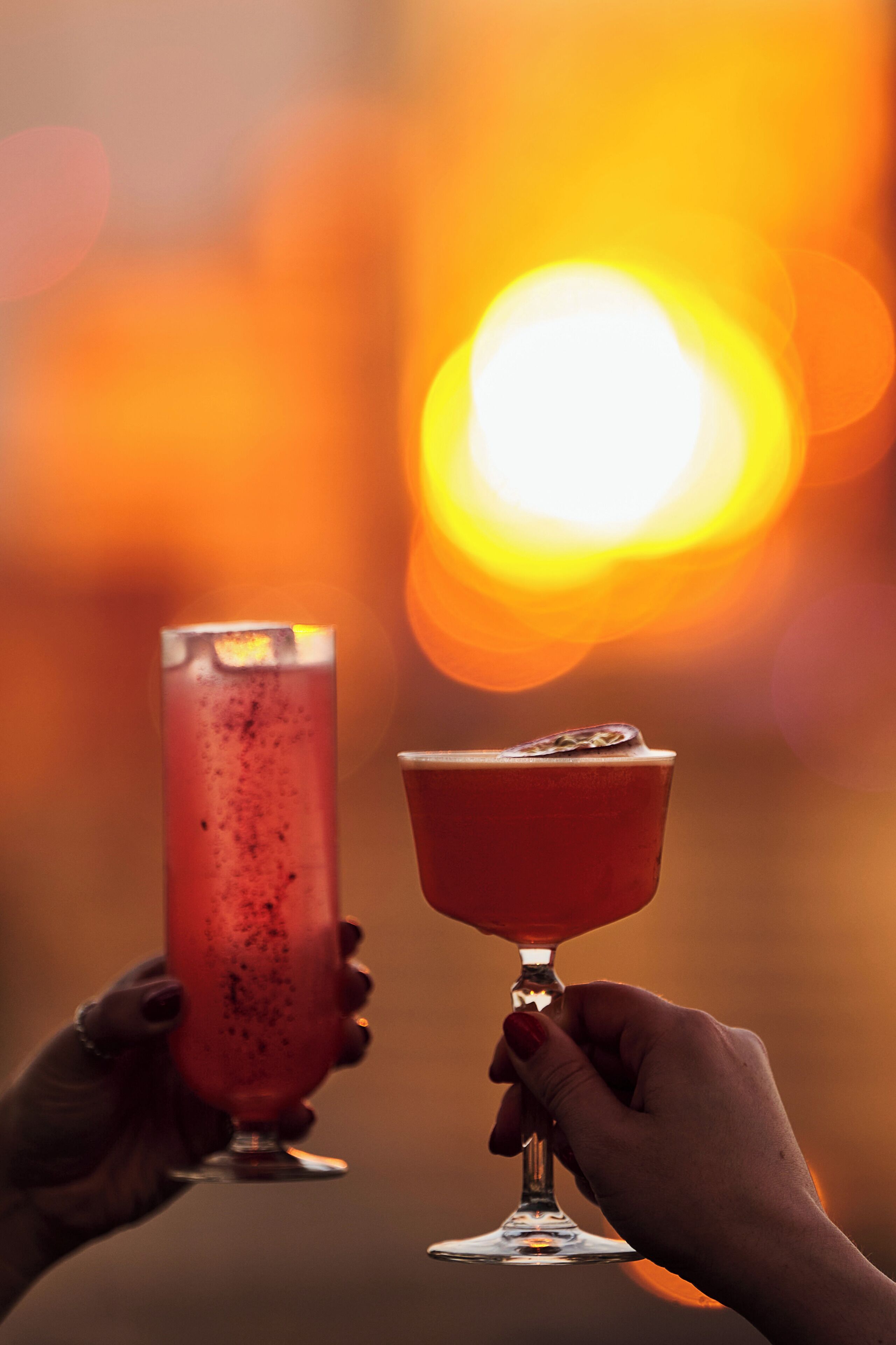 Two hands holding cocktail glasses against a sunset background.