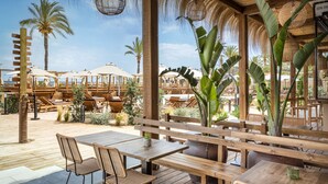 A wooden deck with tables and chairs, surrounded by palm trees and umbrellas.