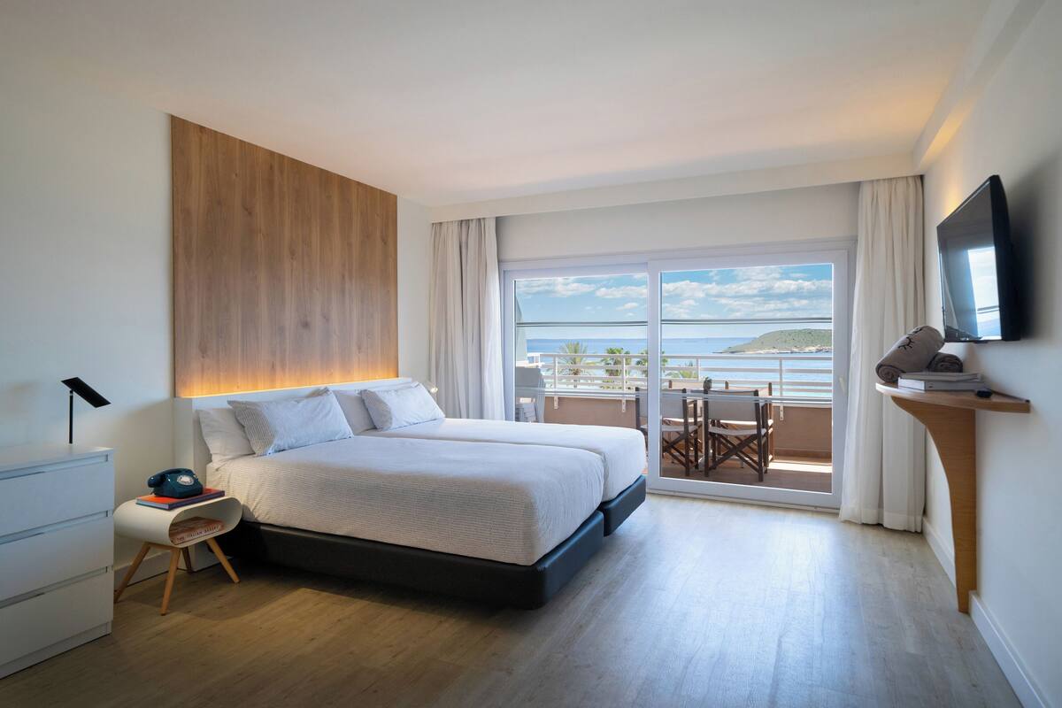 Habitación de hotel moderna con una cama grande, un escritorio, una silla, un televisor y un balcón con vistas al mar.