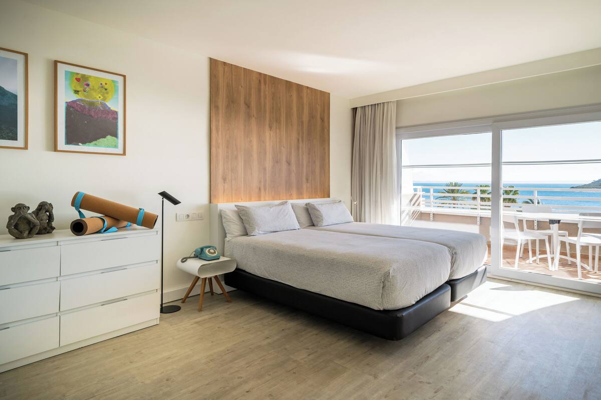 Un dormitorio con cama, escritorio, silla, aparador y balcón con vista al mar.