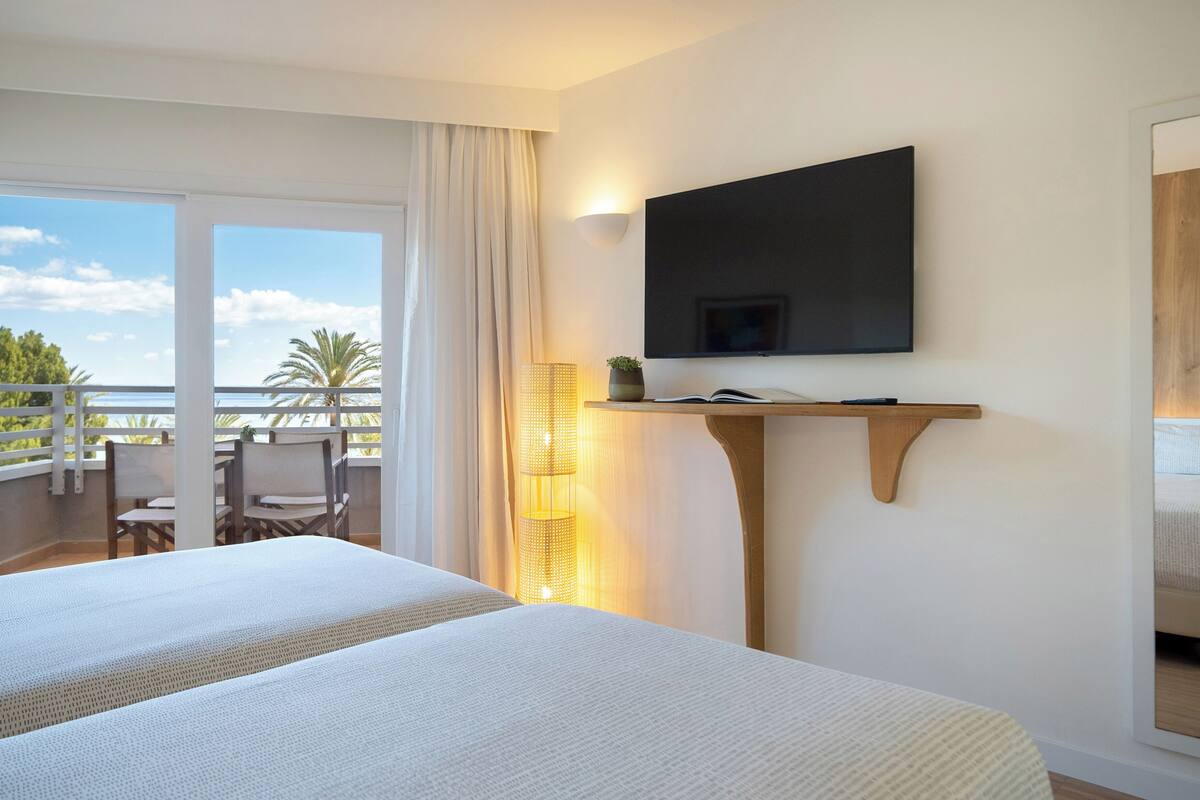 Habitación de hotel con una cama grande, televisión montada en la pared, balcón con sillas y vista al mar.