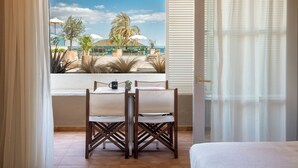 A room with a large window overlooking a beach, a dining table with chairs, and a bed.