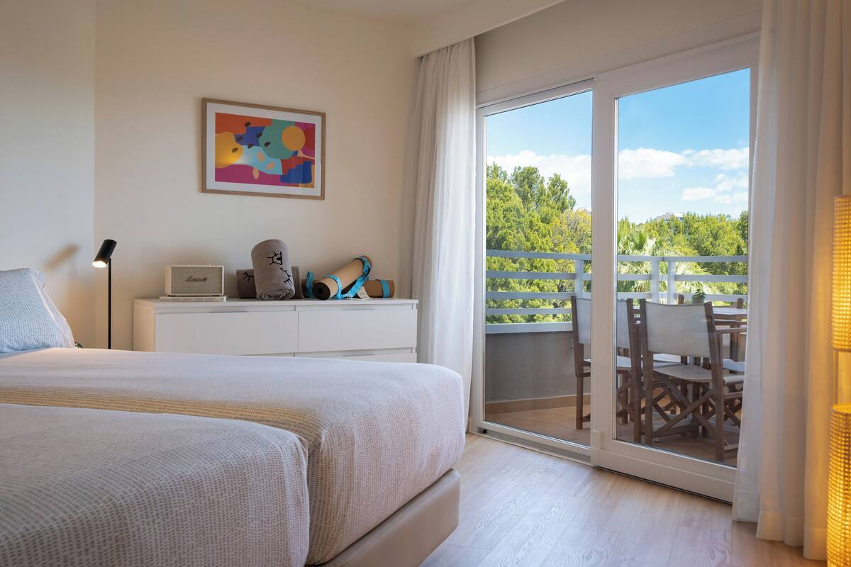 Habitación de hotel con cama, escritorio con lámpara, ventana con vista a follaje y balcón con mobiliario exterior.