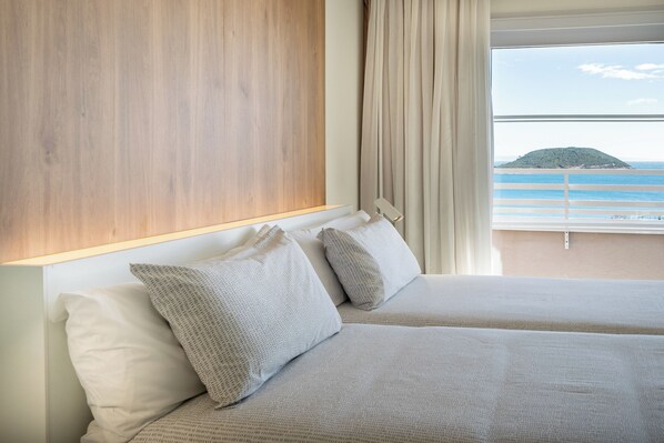 Un cuarto de hotel con cabecera de madera, dos camas con ropa de cama gris, un ventanal con vista al mar y una isla distante.
