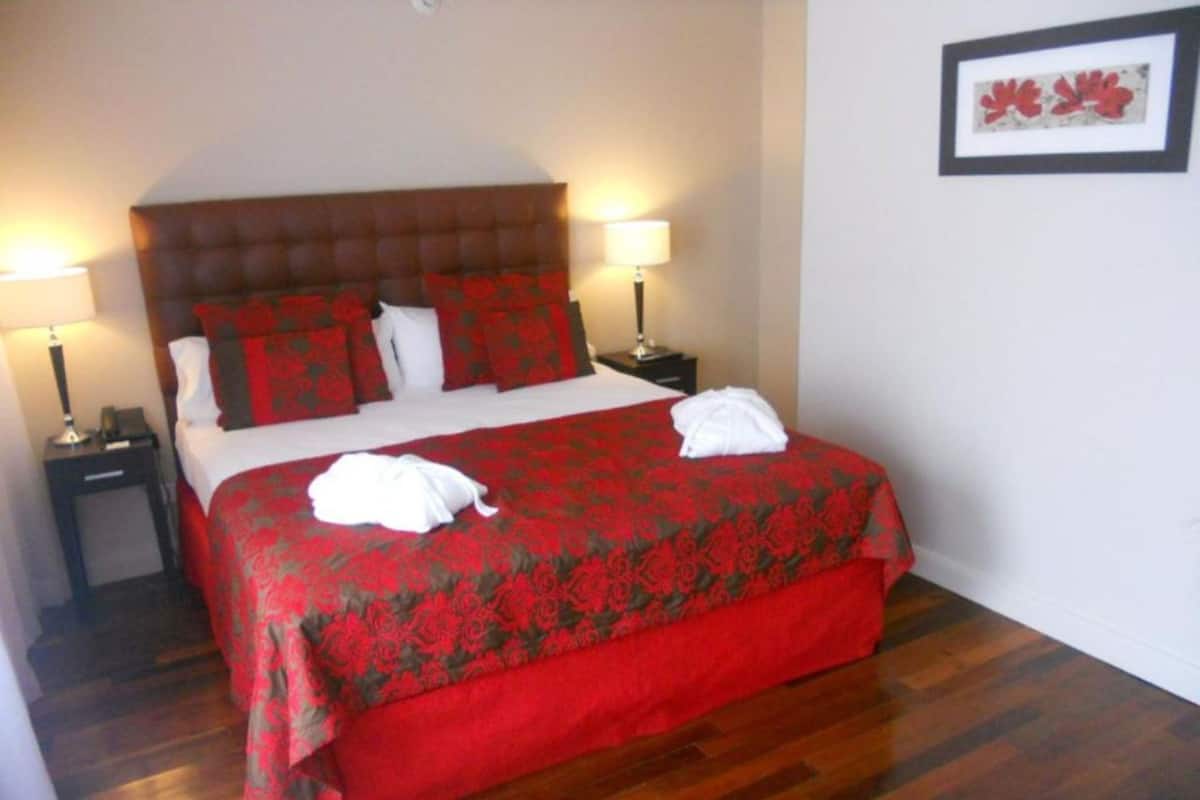 Una cama bien hecha con una colcha de diseño rojo y negro, dos toallas blancas, dos lámparas de noche y un cuadro en la pared.