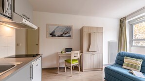 A modern kitchen with a built-in stove, a dining area with a wooden table and a chair, a blue sofa with a striped pillow, and a sailboat painting on the wall. - Lagrange Apart'HOTEL L'Escale Marine (La Rochelle)