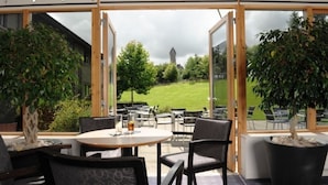 Un espace extérieur aménagé avec des tables et des chaises rondes, de grands pots de plantes et une vue sur un champ verdoyant et un édifice qui se dresse au loin.
