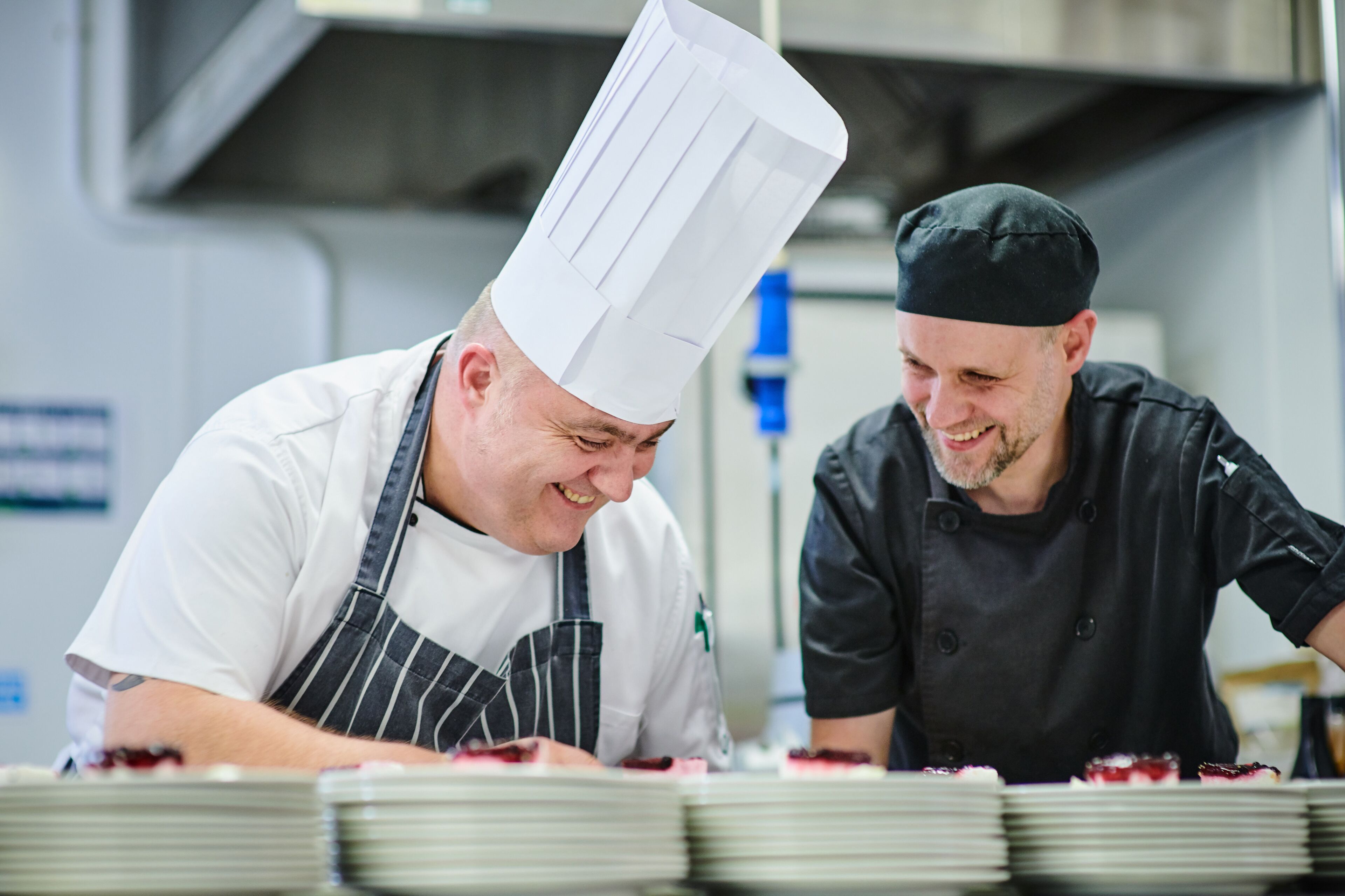 Dos cocineros en una cocina, uno con gorro y mandil blanco, y el otro con uniforme negro.
