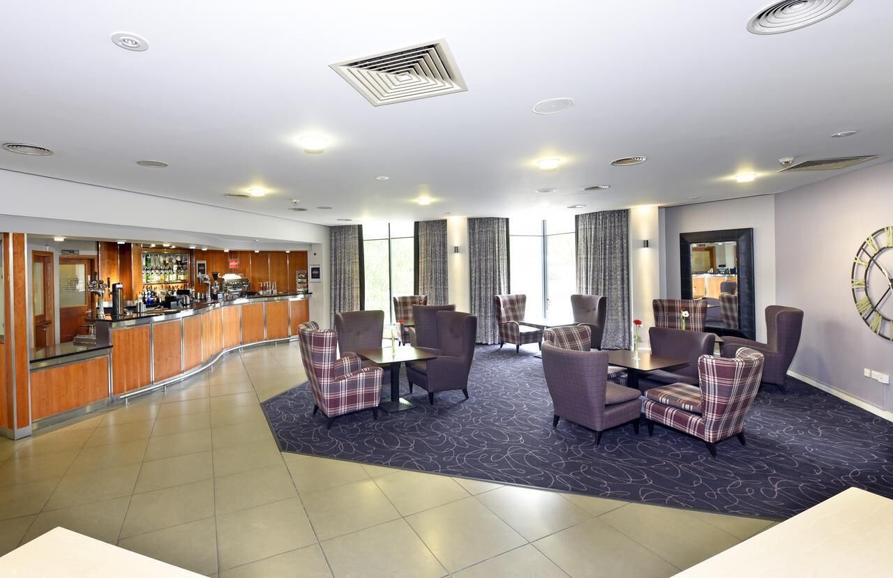 En modern hotellobby med en böjd receptionsdisk, en sittgrupp med fåtöljer och ett bord, stora fönster med gardiner och en klocka på väggen.