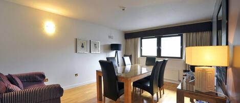A hotel room with a dining table, chairs, a sofa, a lamp, and a window with curtains.