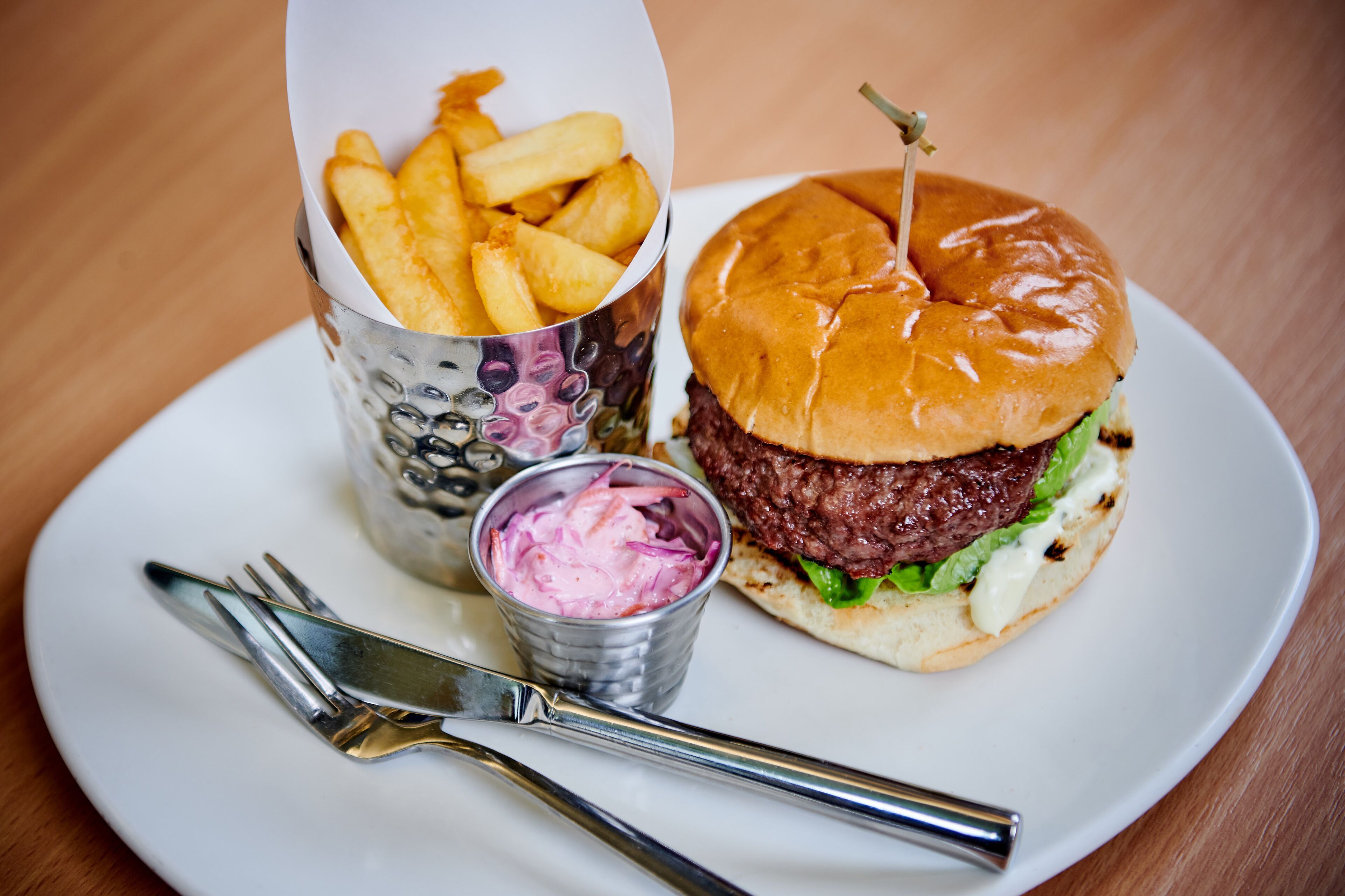 Une assiette avec un hamburger et des frites, un petit bol de salade de chou, et une fourchette et un couteau sur une table en bois.