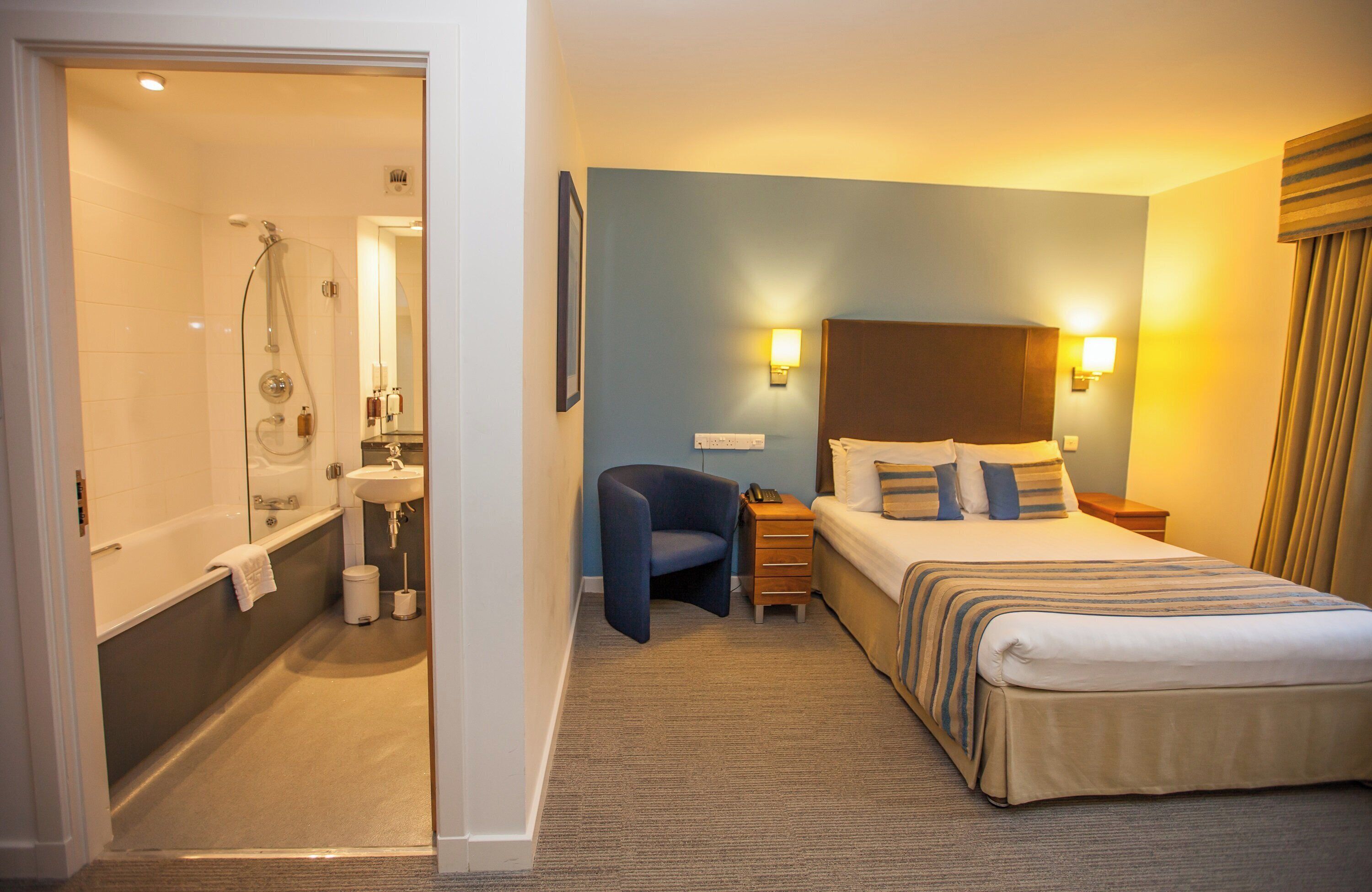 Quarto de hotel com chuveiro, cama com almofadas, mesa de cabeceira e uma cadeira.