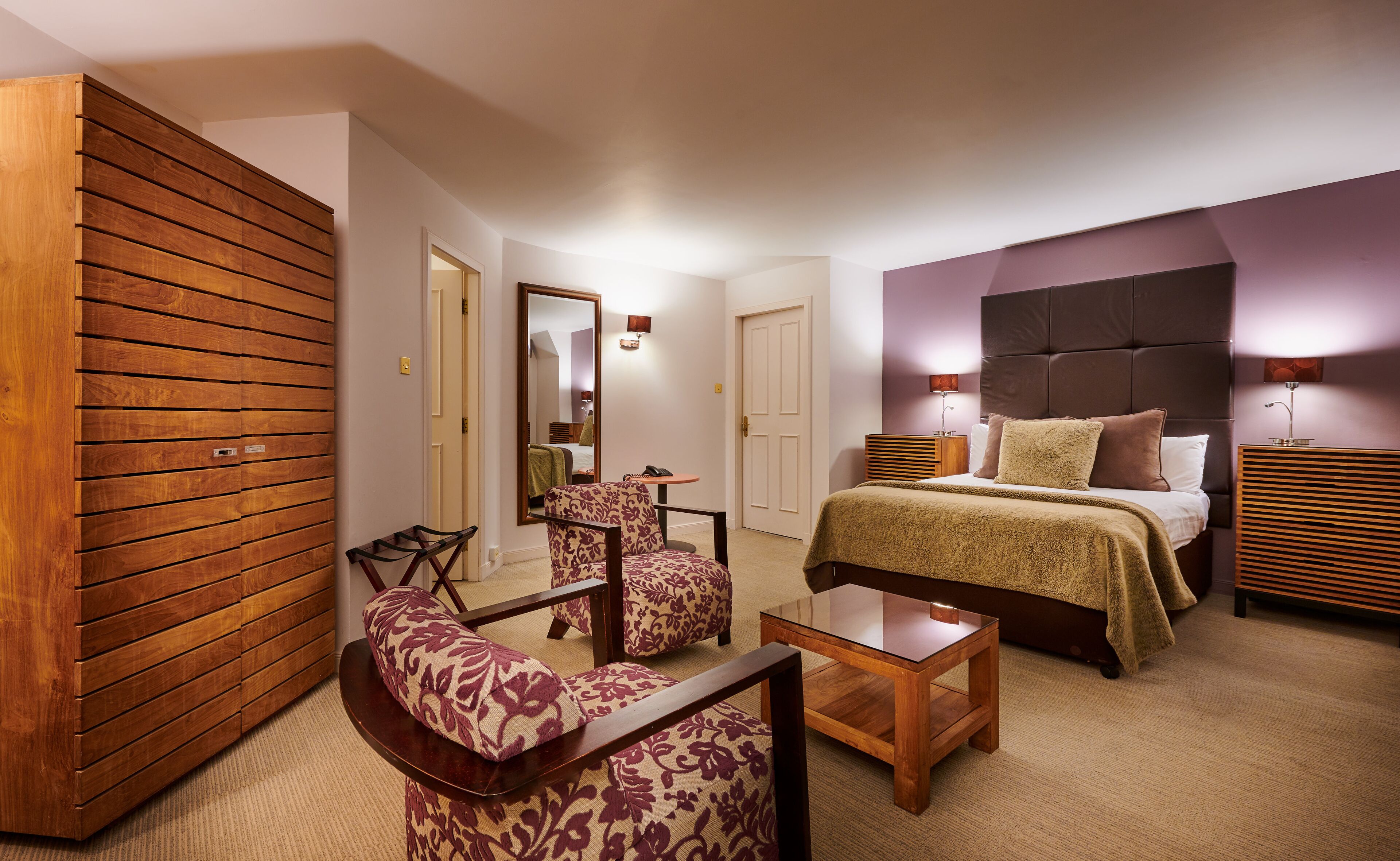 Habitación de hotel con cama, armario de madera, zona de estar con sillas estampadas, una mesita y un espejo.