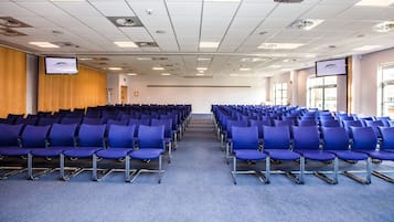 A conference room with rows of blue chairs, a wooden panel wall, and a large screen on the left.