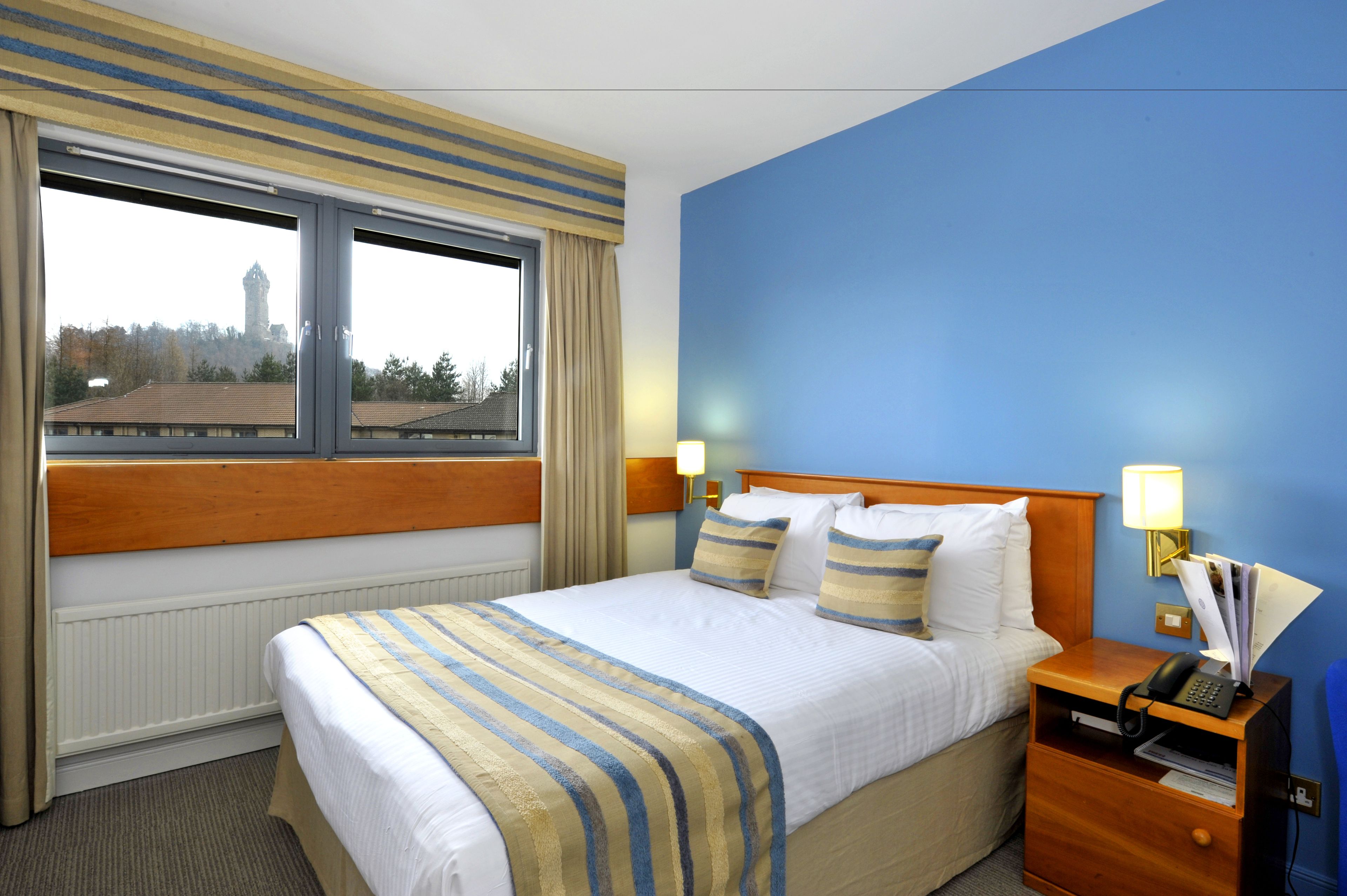 A hotel room with a large bed, a wooden headboard, a nightstand with a lamp, and a view of a building through the window.