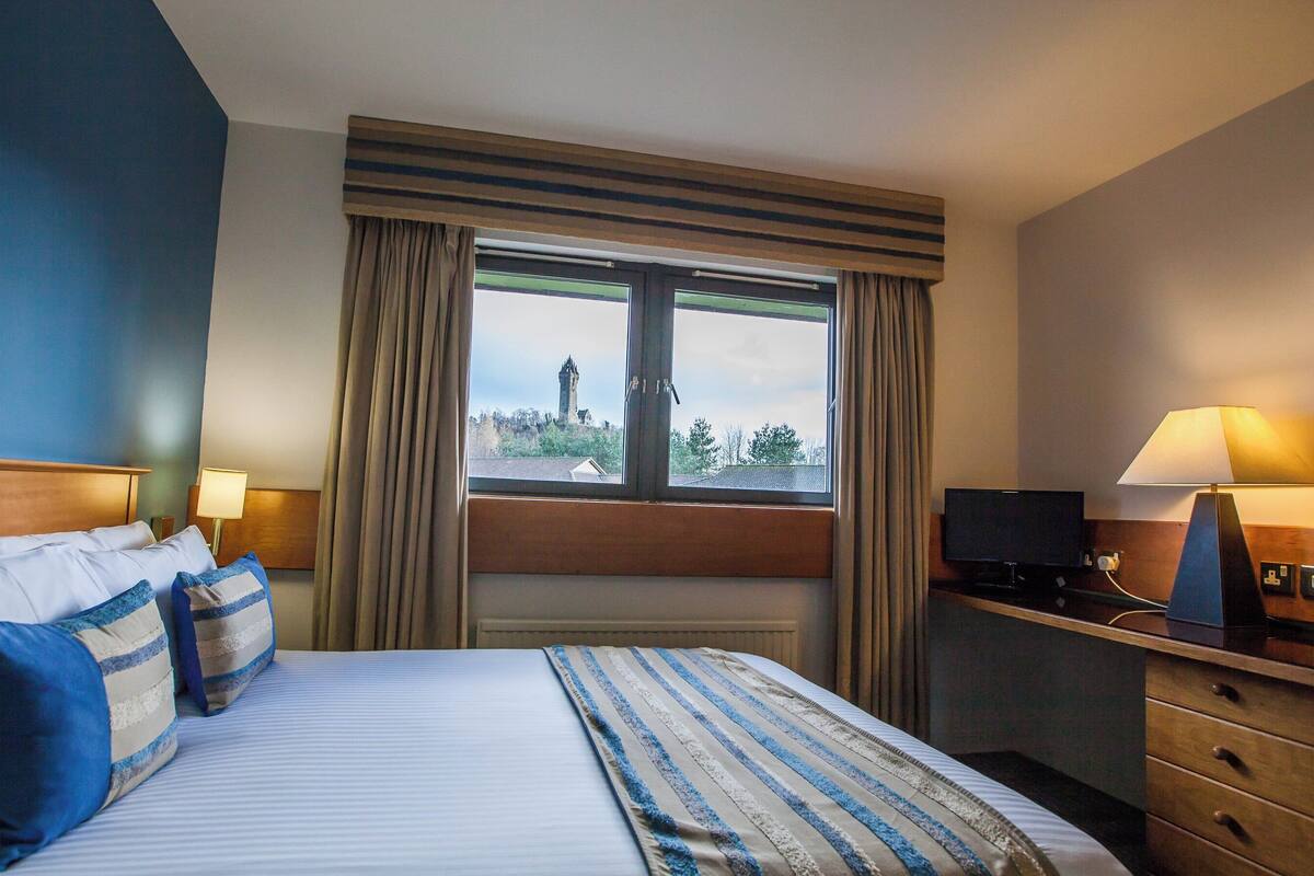 Una habitación de hotel con una cama, dos lámparas de noche, un escritorio con ordenador y una ventana con cortinas.