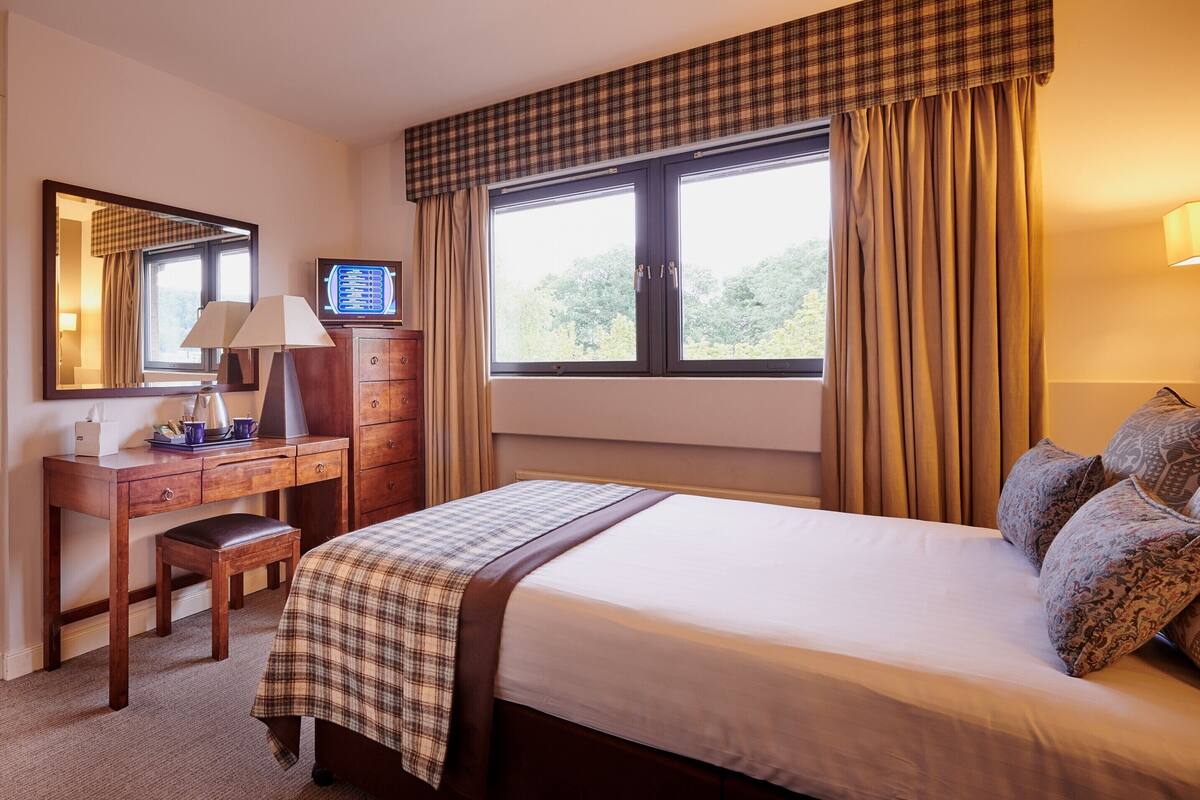 Una habitación de hotel con cama, escritorio con lámpara, televisor y ventana con cortinas.