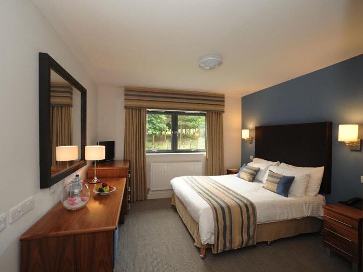A hotel room with a bed, bedside lamps, a mirror, a window with curtains, a wooden dresser, and a small table with a fruit bowl.