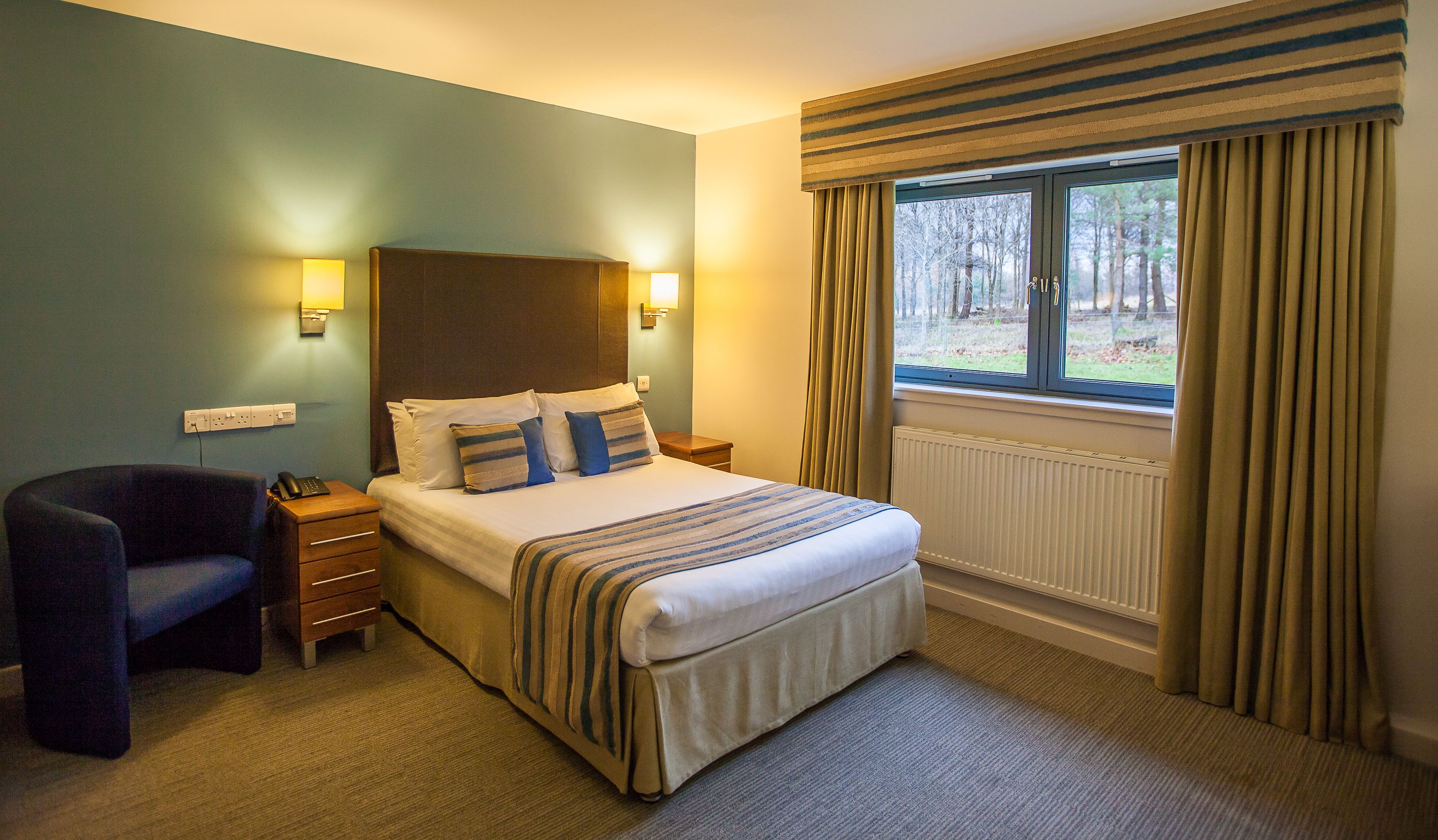 Une chambre d’hôtel avec un lit, des lampes de chevet, une chaise et une fenêtre avec des rideaux.