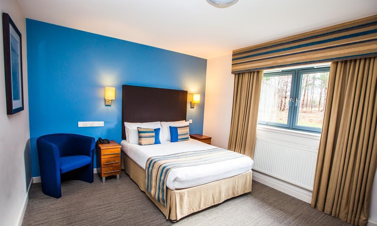 Habitación de hotel con una cama grande, pared azul, ventana con cortinas, escritorio con lámpara y una silla.