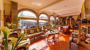 A spacious living room with a sofa set, a dining table, and chairs. There are large windows with arches, a chandelier, and various decorative items. - Fuente de Agua Cusco Hotel (Cusco)
