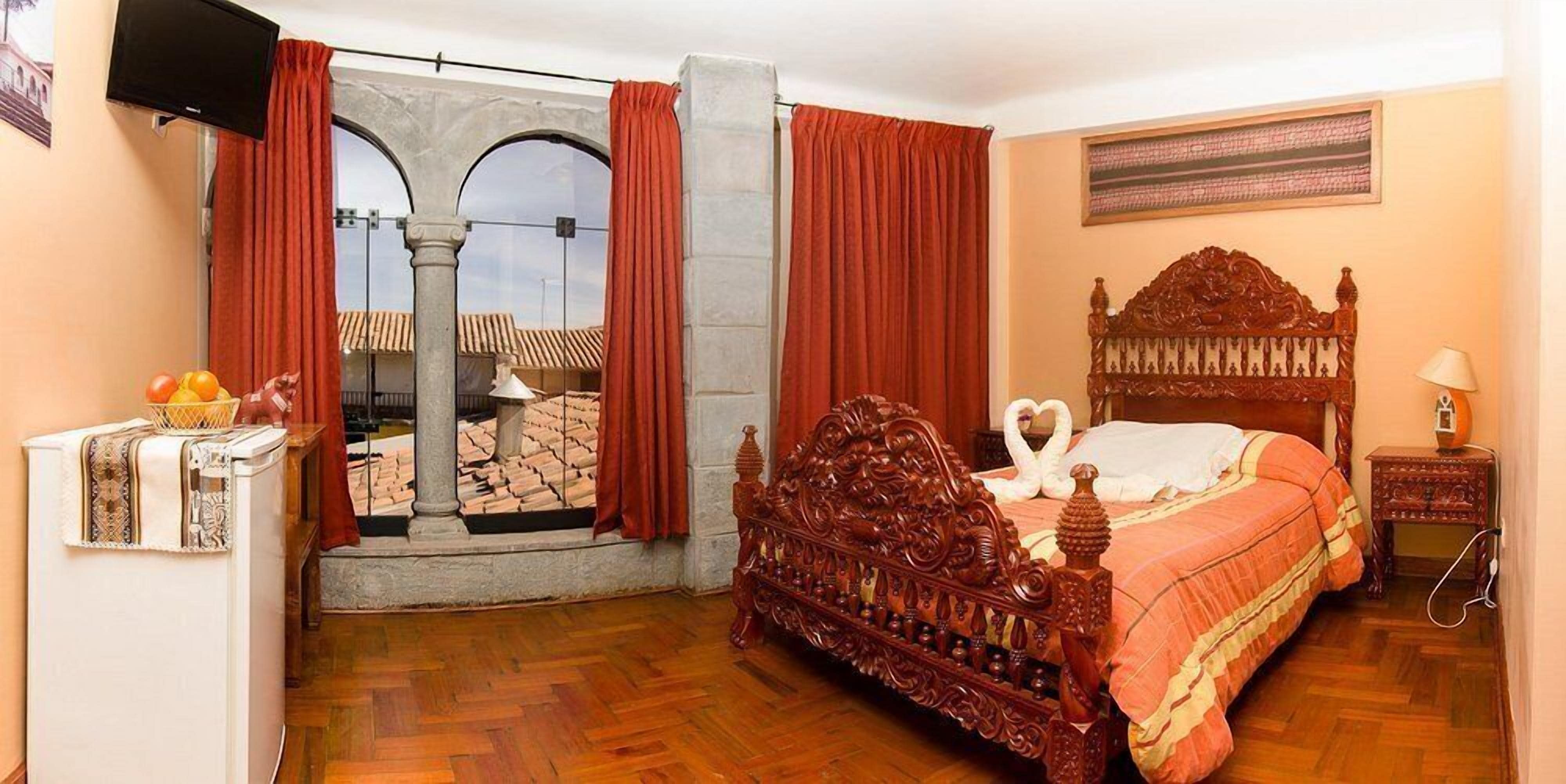 Ein Schlafzimmer mit einem hölzernen Bett, einem Fernseher, einer Obstschale und Blick durch ein Fenster mit roten Vorhängen.
