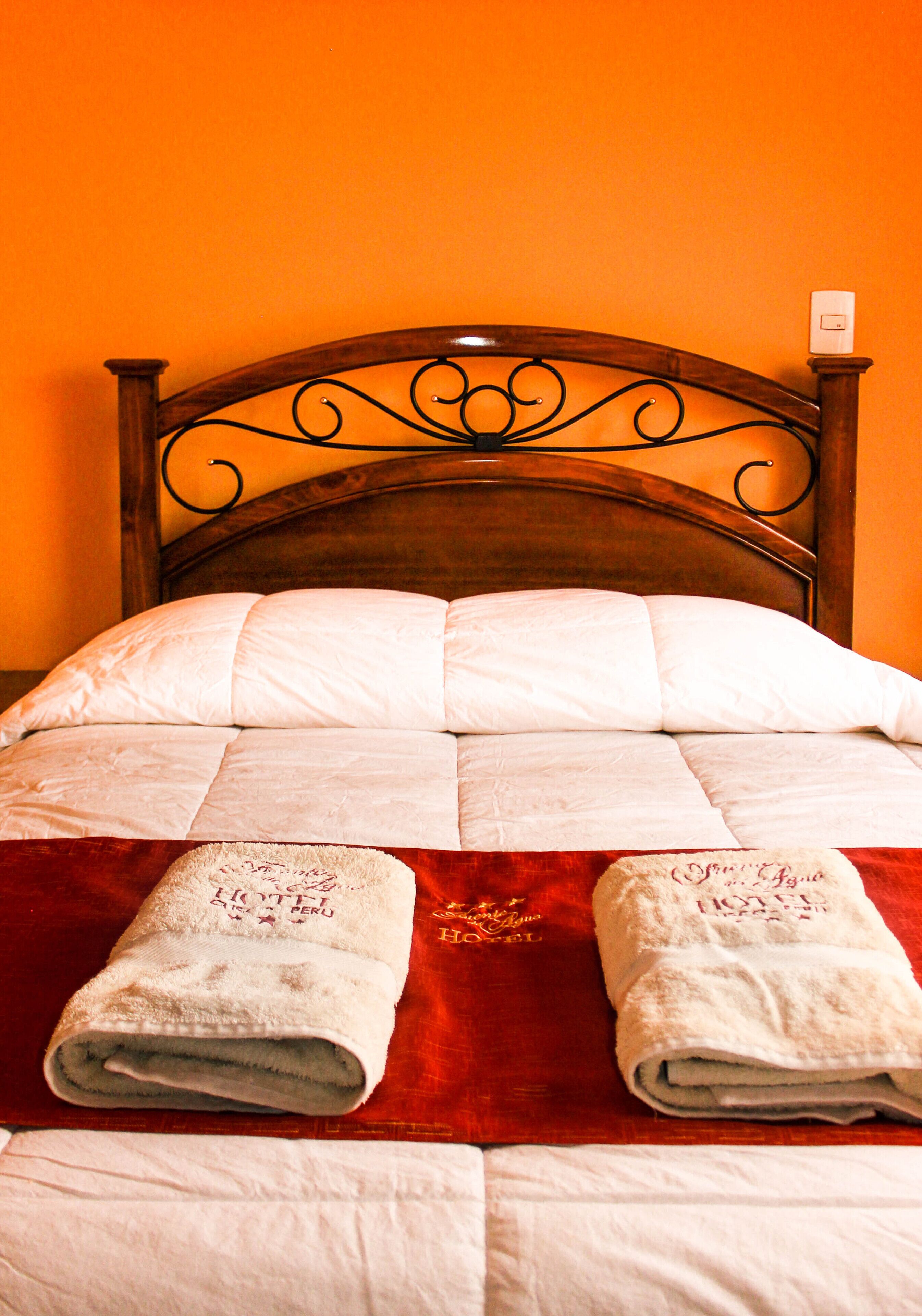 A neatly made bed with a wooden headboard, white linens, and two towels with embroidered text.