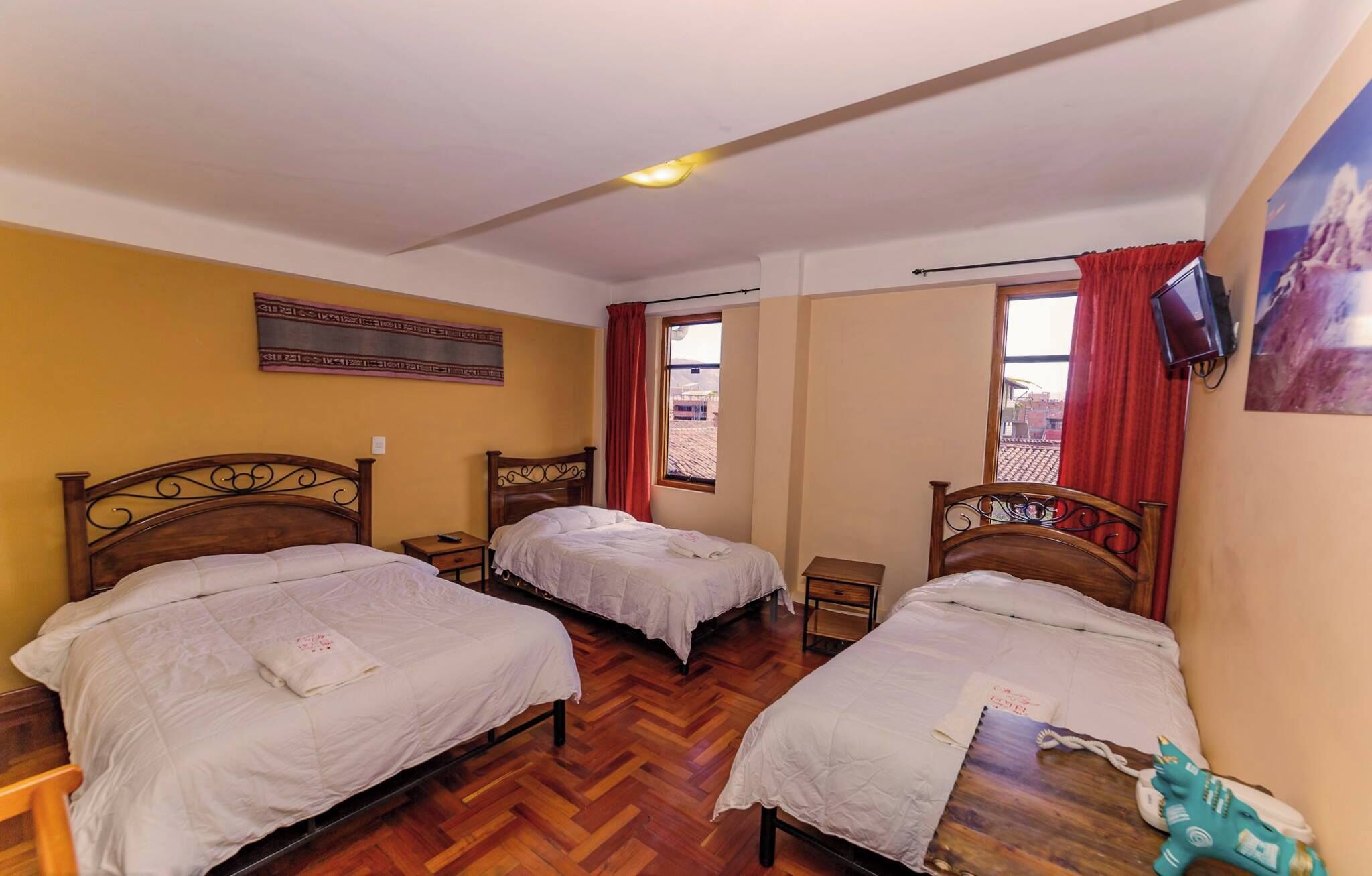 A hotel room with two single beds, wooden headboards, a small bedside table with a lamp, a window with red curtains, and a mounted television.