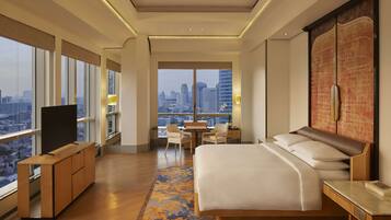 A modern hotel room with a large bed, a desk, and a TV. It features large windows with a city view.