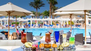 Un bar junto a la piscina con diversas bebidas y frutas, con vistas a una zona de piscina con sombrillas y sillones de descanso.