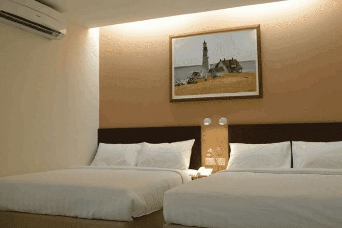 A hotel room with two beds, a nightstand, a wall-mounted lamp, and a framed picture of a lighthouse.
