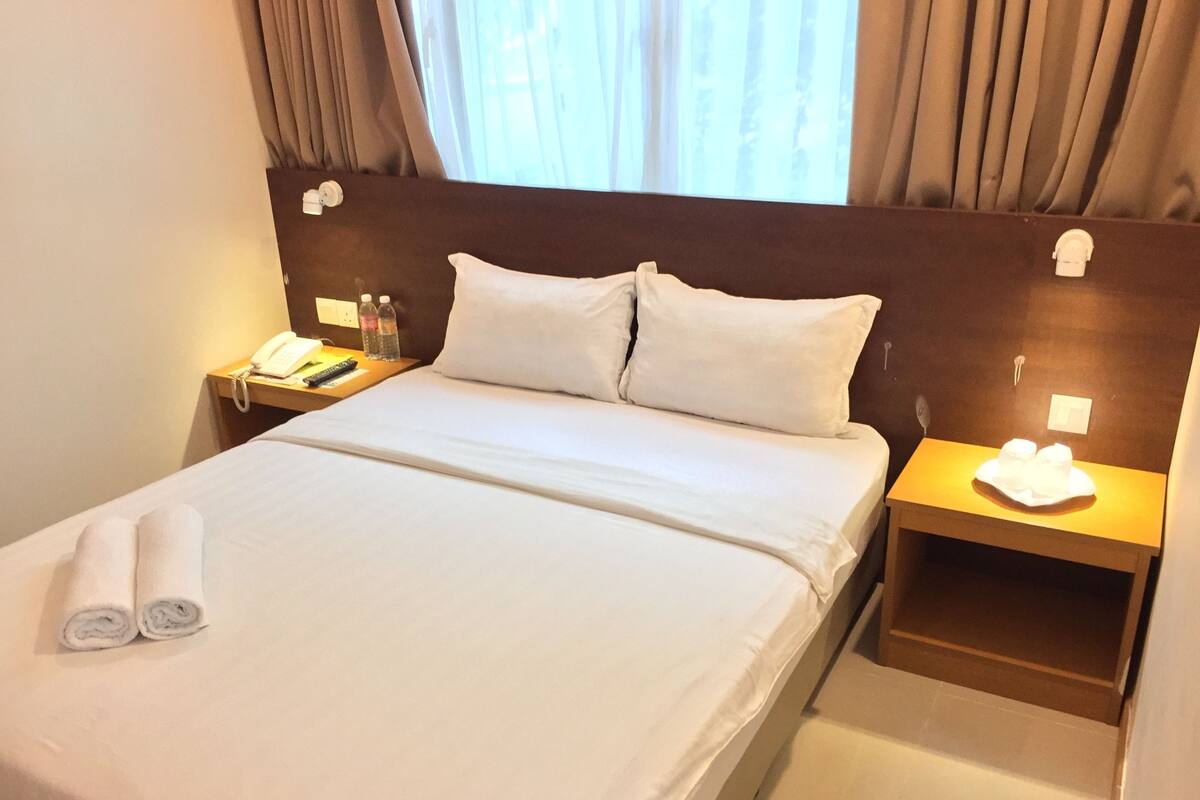 A hotel room with a neatly made bed, a bedside table with a lamp, a phone, and a bottle of water.