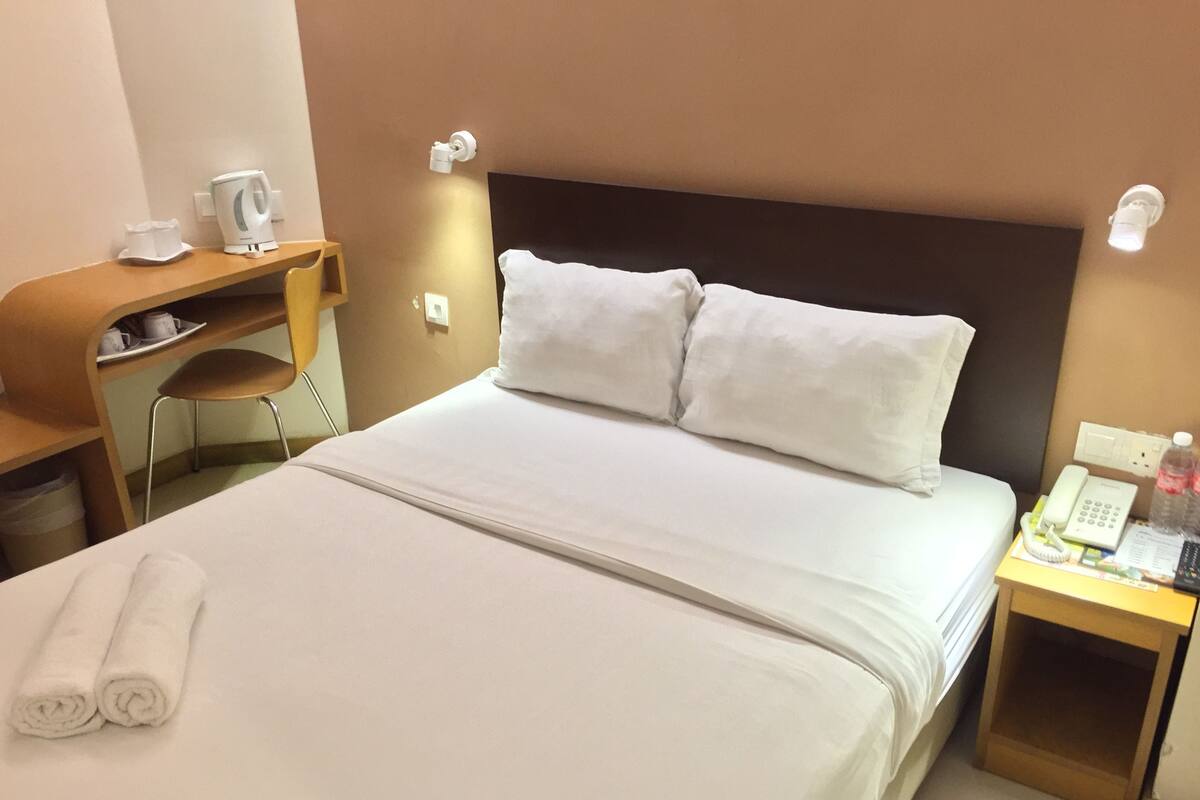 A hotel room with a neatly made bed, a desk with a chair, a telephone, and a small table with a bottle and papers.