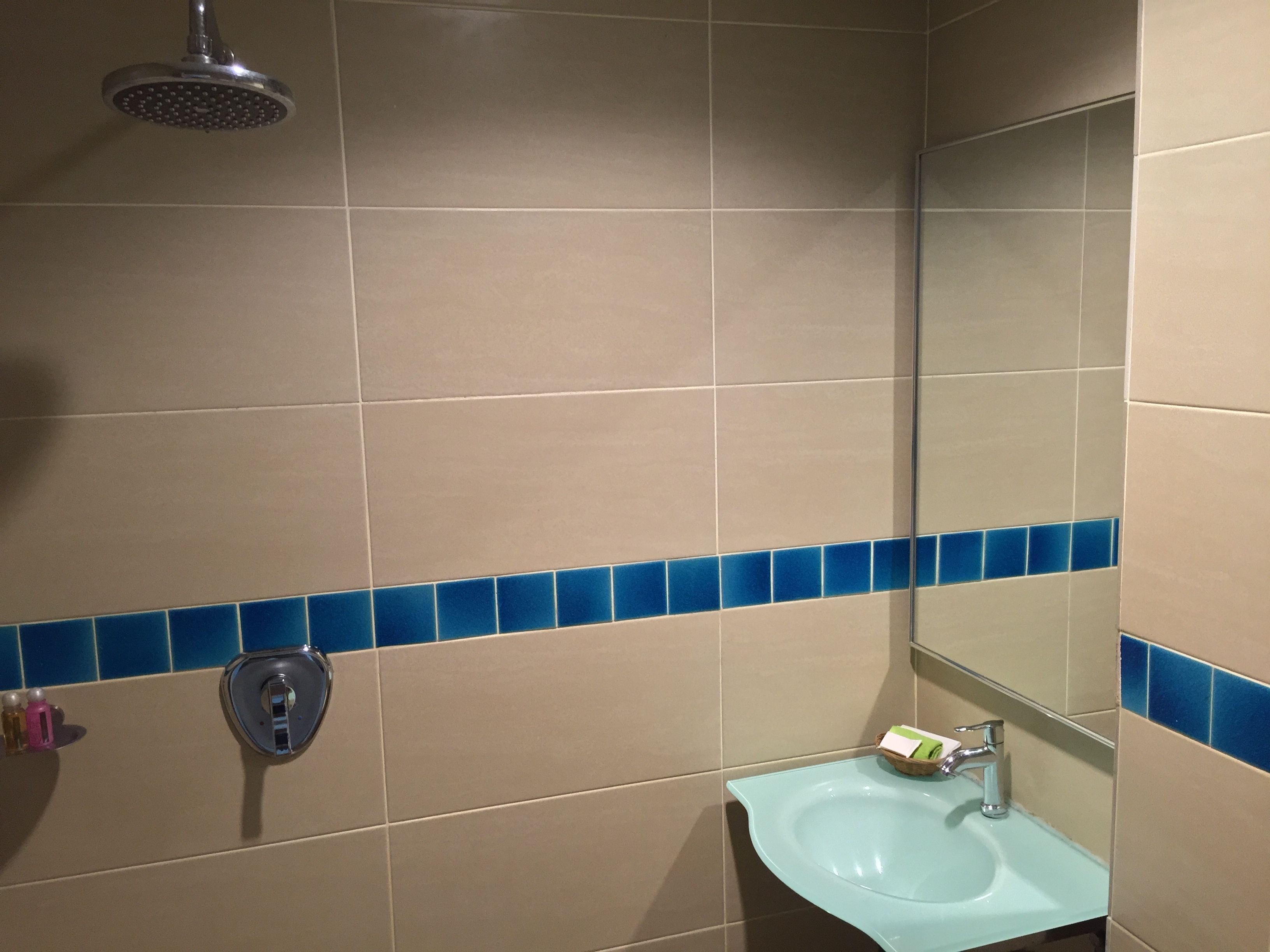 A bathroom with beige tiles, a blue-tiled accent strip, a glass sink, a handheld showerhead, and a mirror.